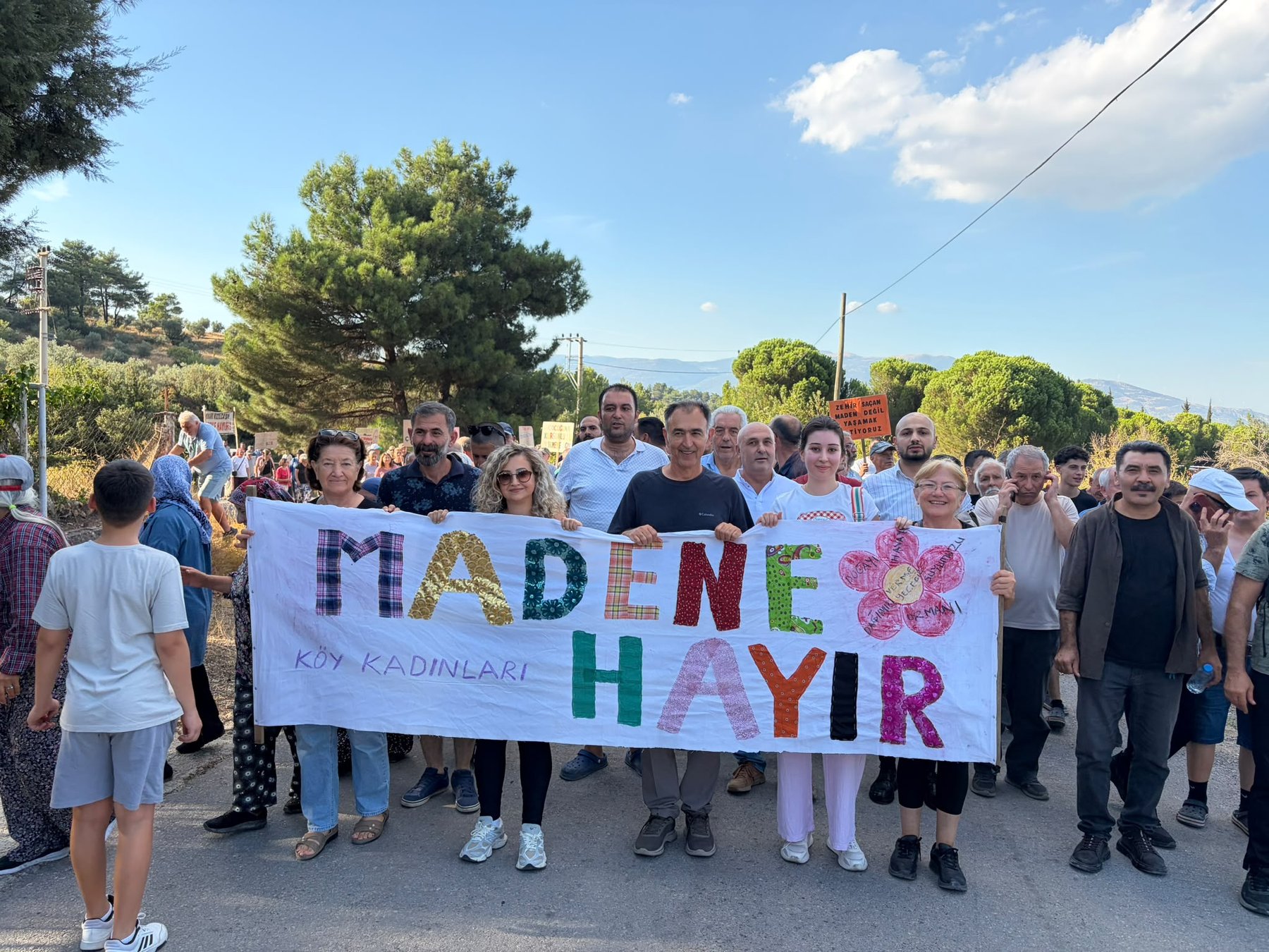 Ağaçlıklı yolda Madene Hayır pankartını taşıyan yürüyüş katılımcıları ve arkalarındaki kalabalık